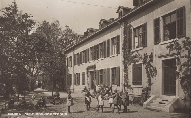 11 spielende Kinder vor dem Missionskinderhaus in Basel. Einige sitzen im Schatten, andere machen einen Spielkreis.