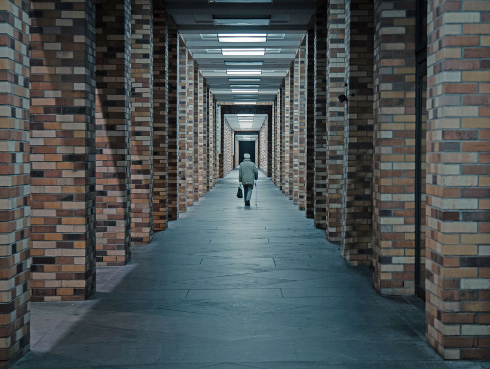 Eine ältere Frau mit Stock läuft alleine einen Gang mit vielen Pfeilern entlang. Mit den Lichtern an der Decke entsteht der Eindruck eines Tunnels.