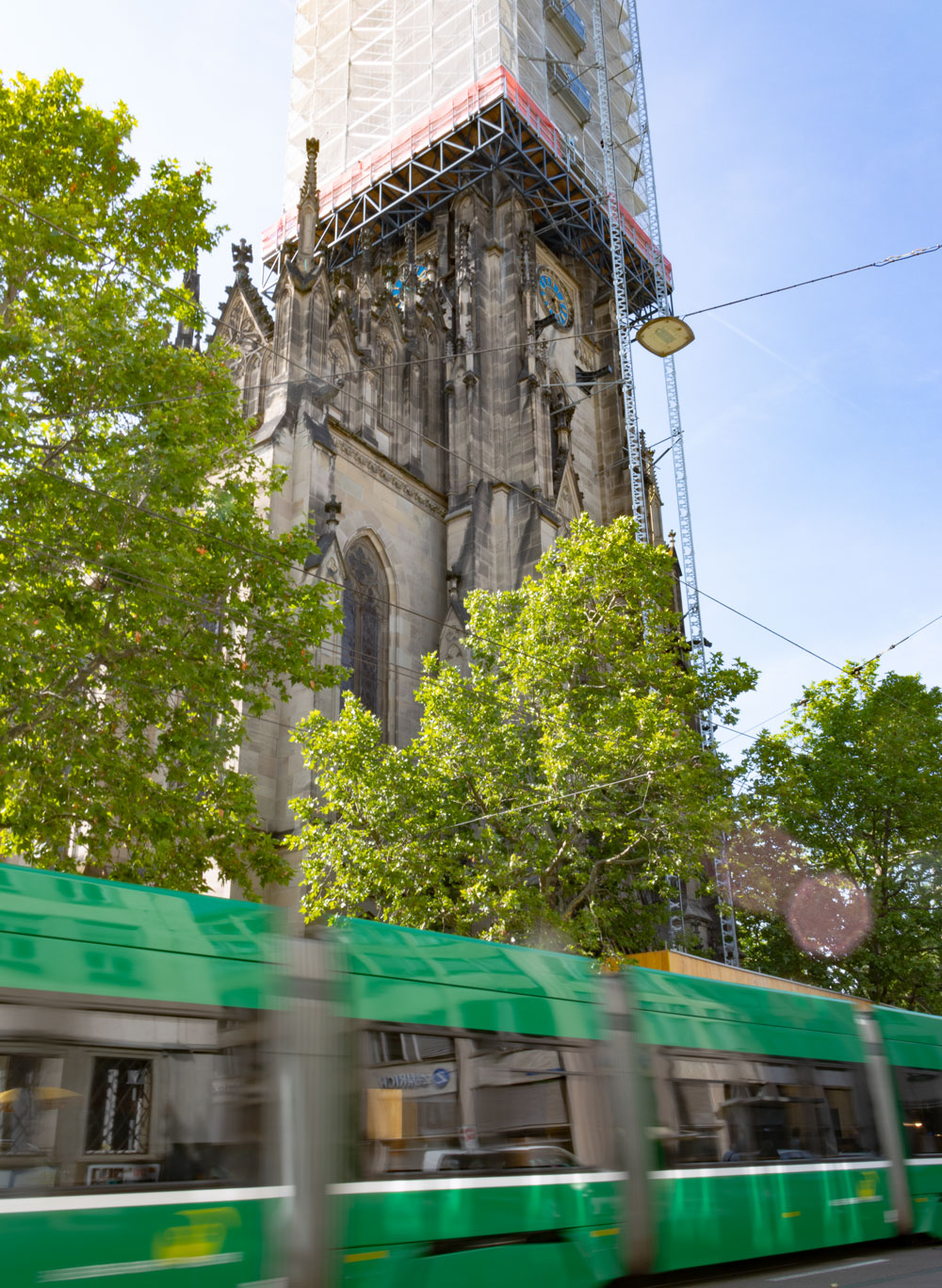 Ein grünes Basler Tram fährt dicht vor dem Eingang der Elisabethenkirche durch. Der Turm ist im Gerüst, die Strassenbahn verschwommen von der Geschwindigkeit.