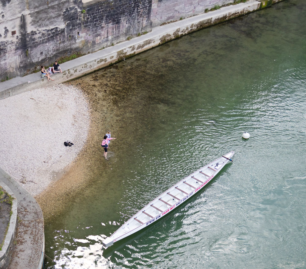 Menschen sitzen am Rheinufer oder stehen barfuss im Wasser neben einem Drachenboot.